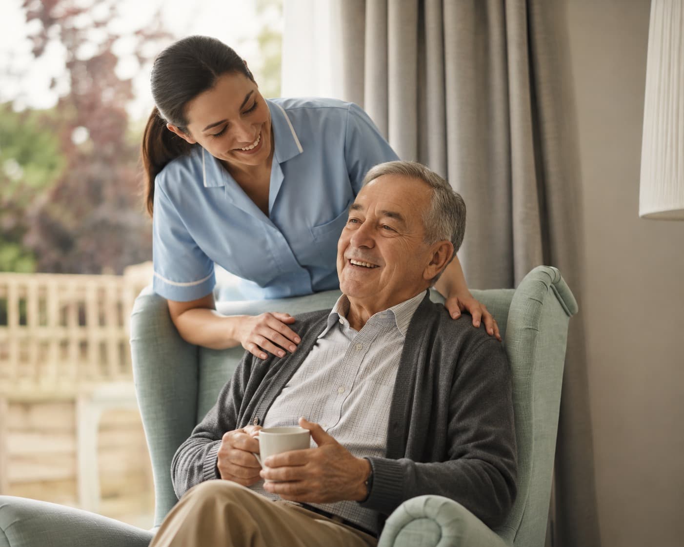 A nurse leaning over an armchair to talk with an older man holding a cup of tea.